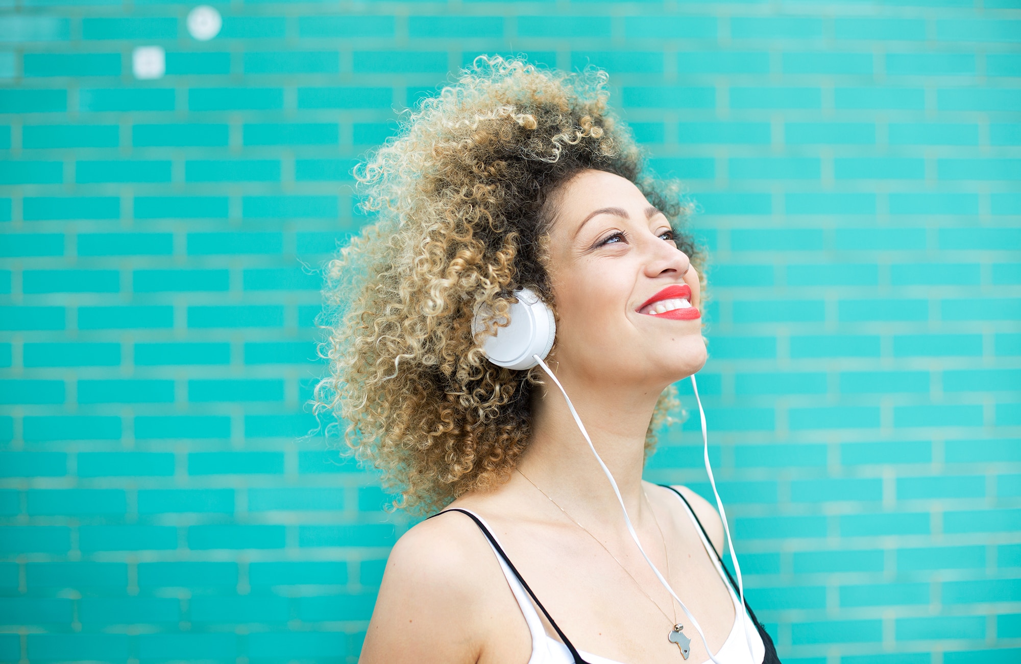 Young woman with a blonde afro and wearing bright red lipstick smiles and listens to music with headphones.