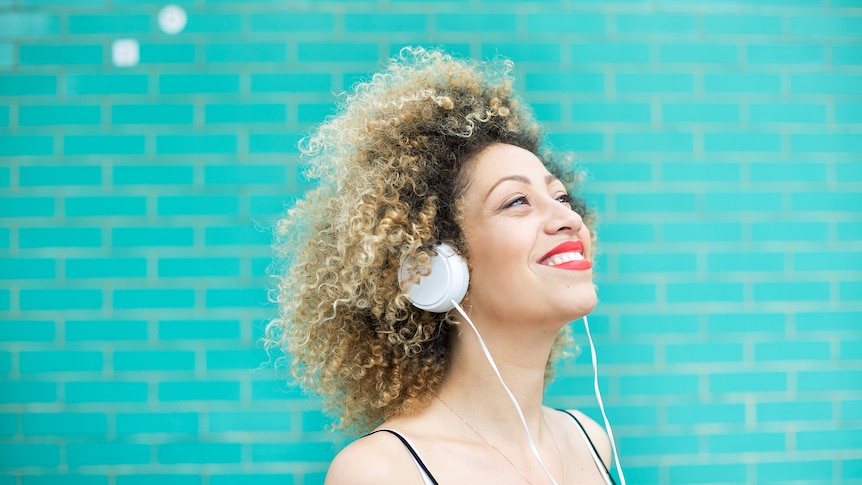 Young woman with a blonde afro and wearing bright red lipstick smiles and listens to music with headphones.