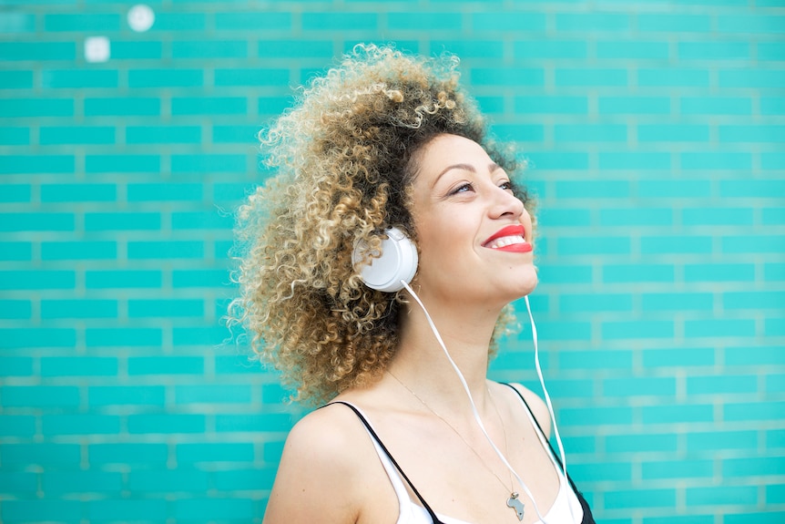 Young woman with a blonde afro and wearing bright red lipstick smiles and listens to music with headphones.