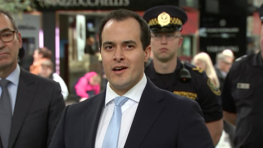 A dark-haired man wearing a suit stands in front of a police officer.
