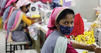 Female Bangladeshi garment maker sewing clothes and wearing a face mask.
