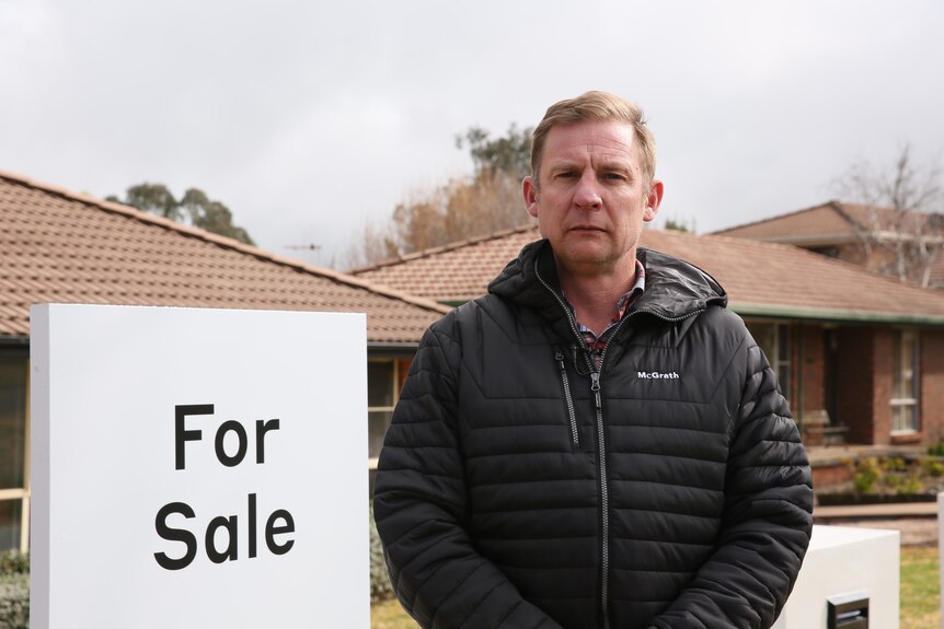 Man in a black puffer jacket standing in front of a 'for sale' sign