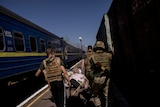 An older man is carried on a stretcher past a train by men wearing military helmets and vests.