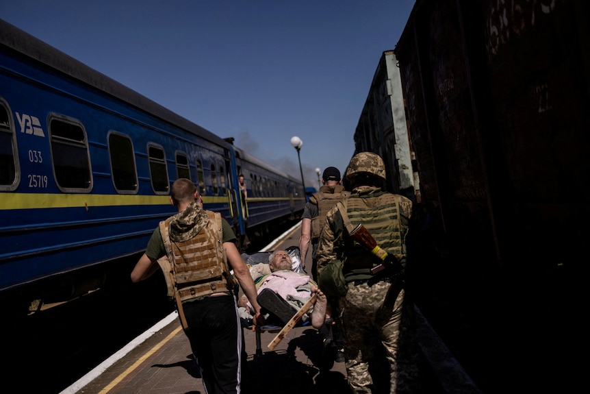 An older man is carried on a stretcher past a train by men wearing military helmets and vests.