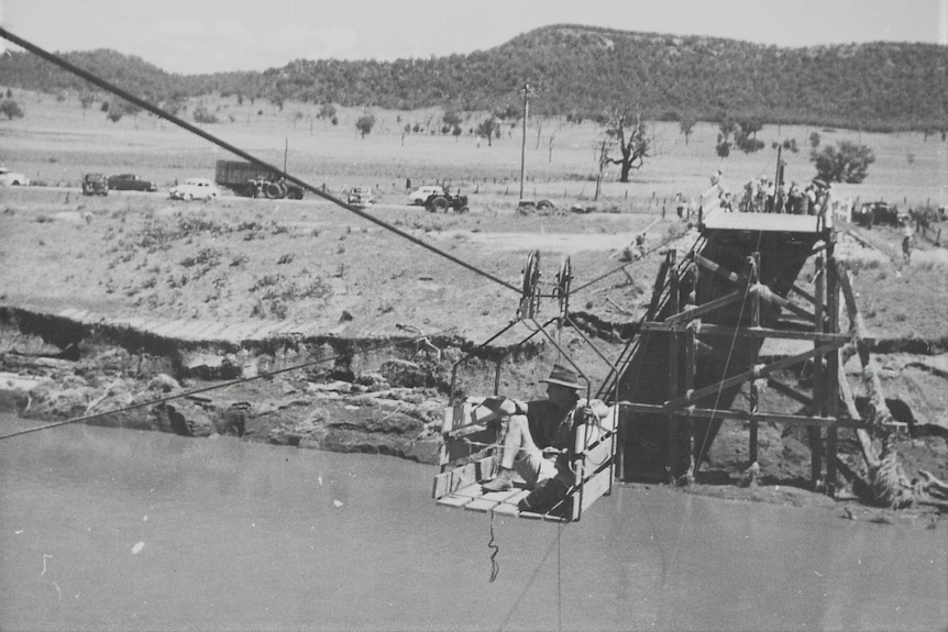 Old photo in black and white showing a man in hat crossing a wide river in a flying fox carriage