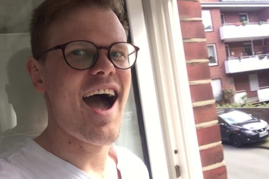 Pascal Herington in white t-shirt and dark rimmed glasses standing at an open window looking out onto a street