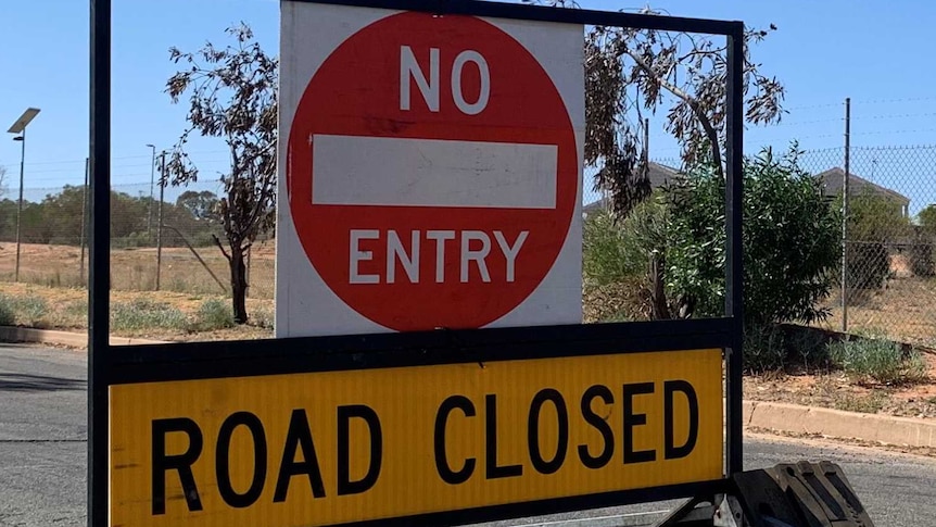 "Road closed", "no entry" sign sits on a street in front of a fenced community and a big blue sky.
