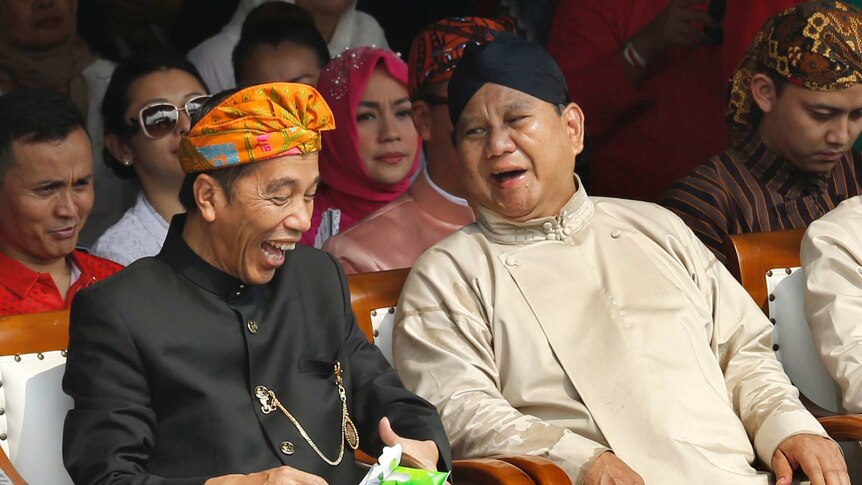 Indonesian President Joko Widodo and Prabowo Subianto laugh as they sit next to each other in a crowd.