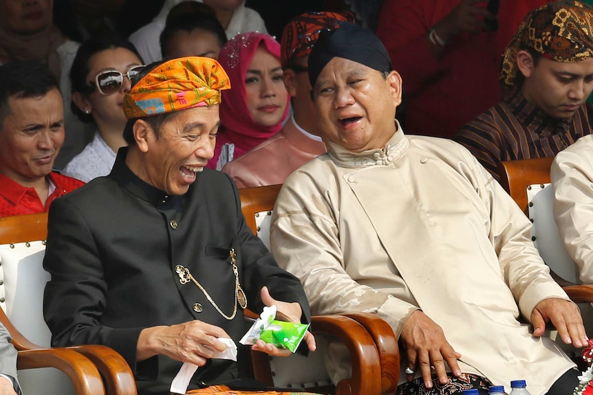 Indonesian President Joko Widodo and Prabowo Subianto laugh as they sit next to each other in a crowd.