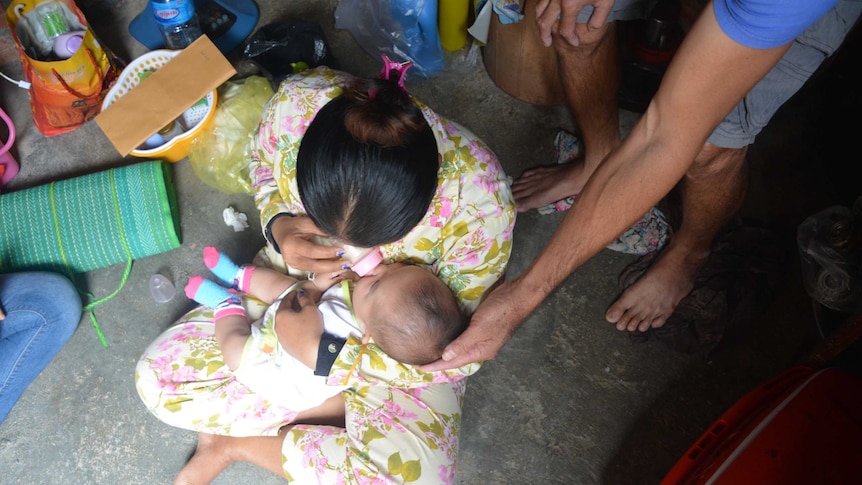 Father cradles baby's head in hand as mother sits on concrete floor with plastic baskets, fan, and gives baby a bottle.