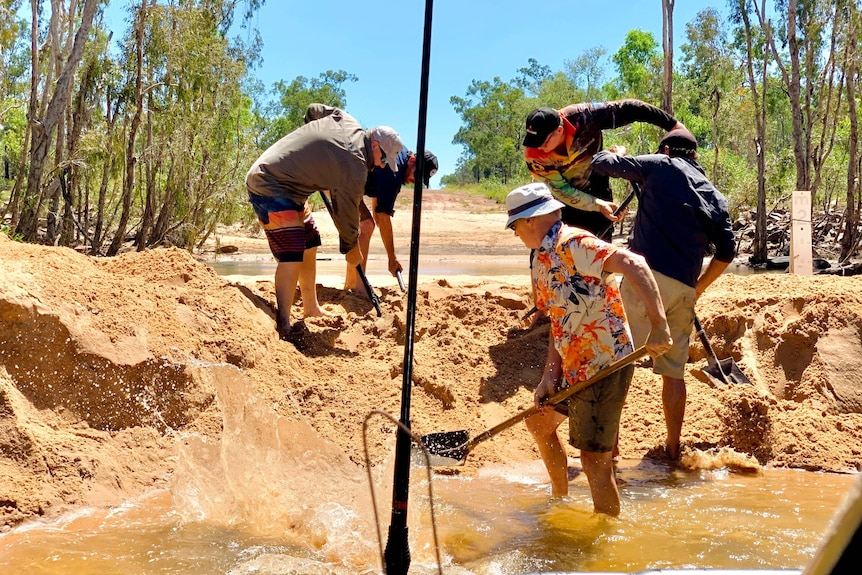 Four men use shovels to dig out some sand at a watercourse.