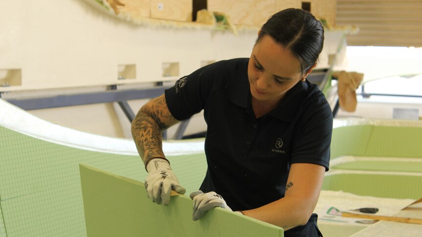 A woman with black hair in navy polo, wearing white gloves, sanding a board