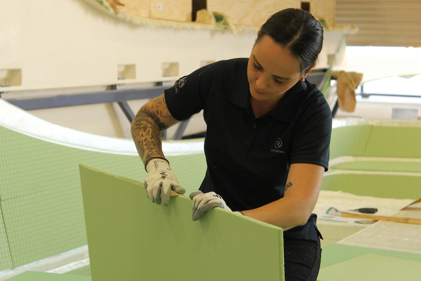 A woman with black hair in navy polo, wearing white gloves, sanding a board