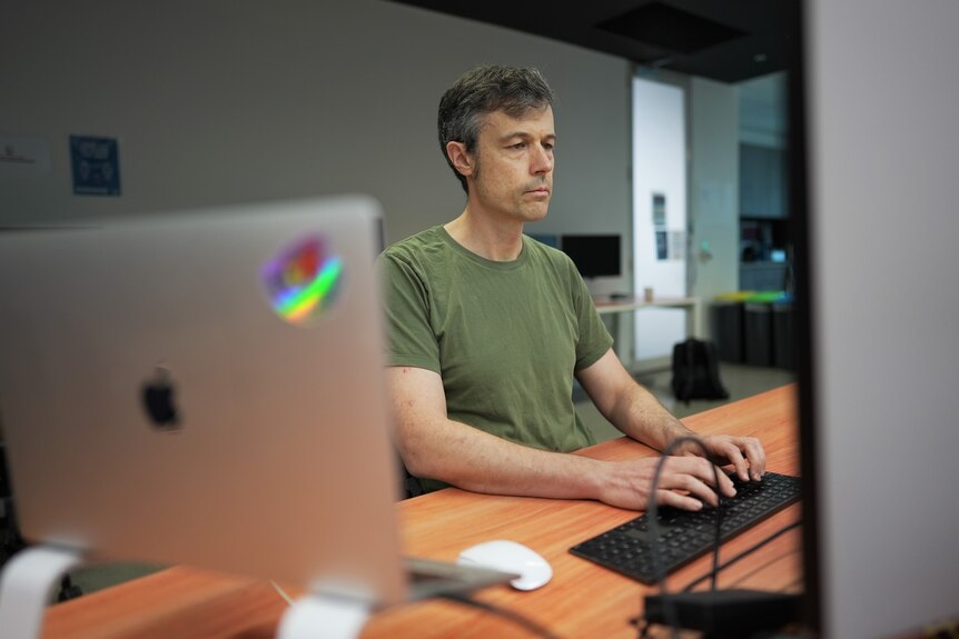 Jamie Twiss, wearing a green t-shirt, sits at a desk typing on a black keyboard. An Apple monitor is seen in the foreground.
