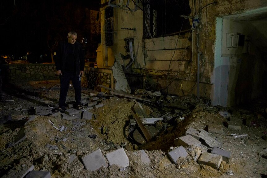 A man looks down at a small crater in the ground next to a darkened, damaged building.