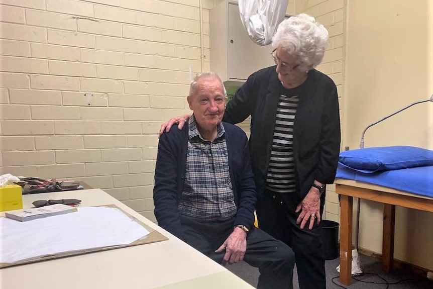 An older man sits down at an office desk, while a woman stands next to him with her arm around him