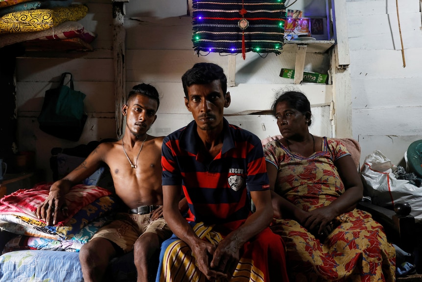 一个斯里兰卡家庭坐在木屋的床上，他们的财物放在临时架子上。