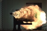 A projectile shot from a railgun fires with a large fiery cloud behind it from a right-angle metal mount.