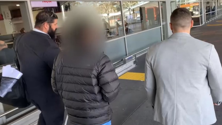 A man with his face blurred is led away by two detectives