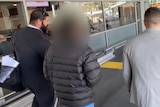 A man with his face blurred is led away by two detectives