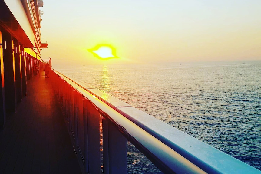 The sun sets over the ocean with the balustrade of a cruise ship in the foreground.
