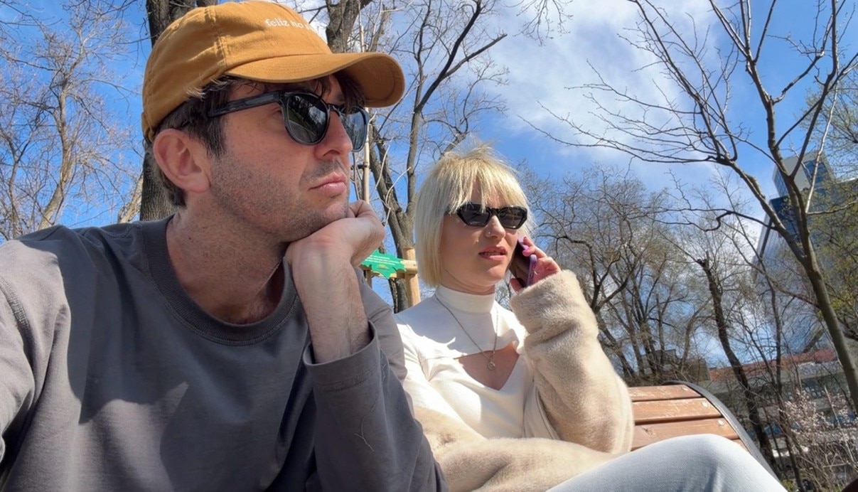 Elliot and Tatiana sit on a park bench while Tatiana speaks on the phone.