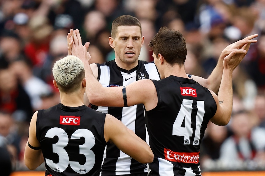 Three Collingwood AFL players embrace as they celebrate a goal.