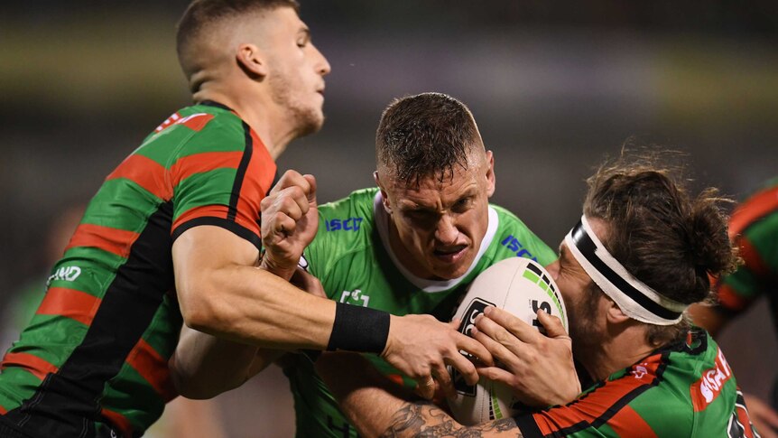 Jack Wighton, in green, centre, is tackled by two players wearing red and green striped jerseys, one on each side.