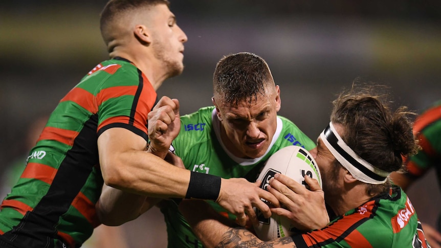Jack Wighton, in green, centre, is tackled by two players wearing red and green striped jerseys, one on each side.