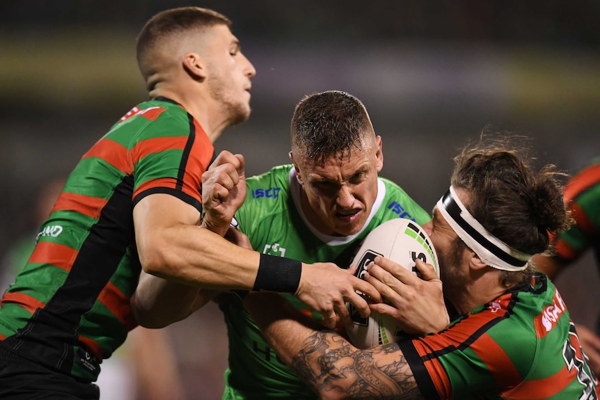 Jack Wighton, in green, centre, is tackled by two players wearing red and green striped jerseys, one on each side.