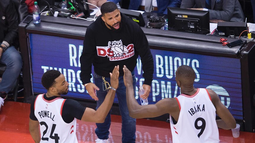 Drake stands just to the side of a basketball court, shouting congratulates towards two Toronto players