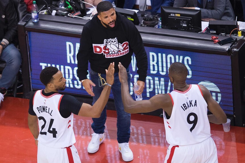 Drake stands just to the side of a basketball court, shouting congratulates towards two Toronto players
