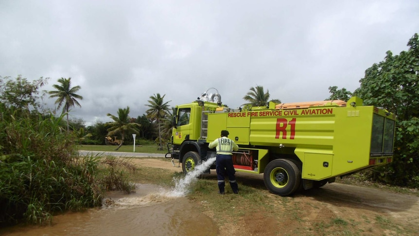 In a tropical landscape, a bright neon yellow firetruck releases water into a brown puddle as palm trees are on the horizon.