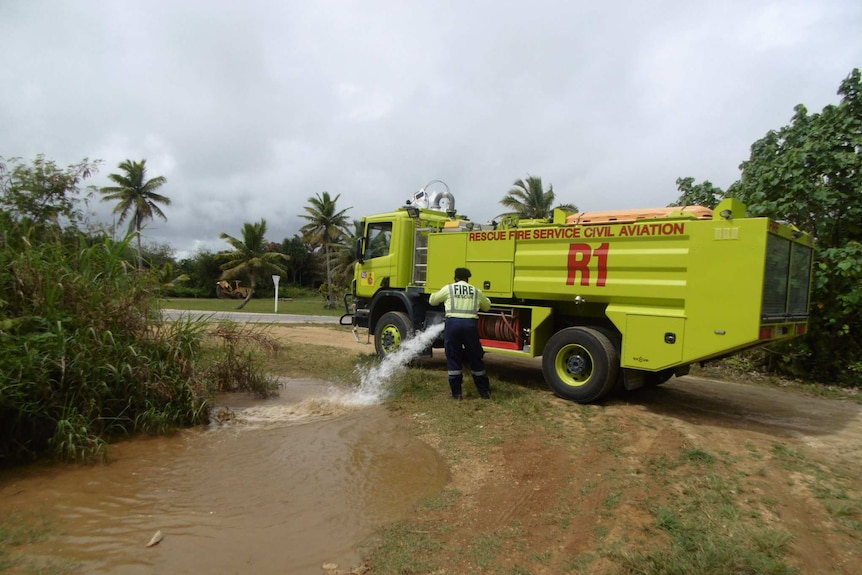 In a tropical landscape, a bright neon yellow firetruck releases water into a brown puddle as palm trees are on the horizon.