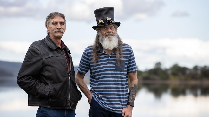 Two middle-aged men in jeans, one wearing a hat, stand in front of a bay