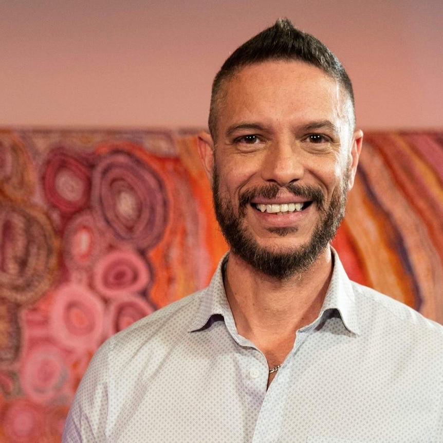 An Aboriginal man in his 40s standing in front of an Aboriginal line and dot painting and smiling.