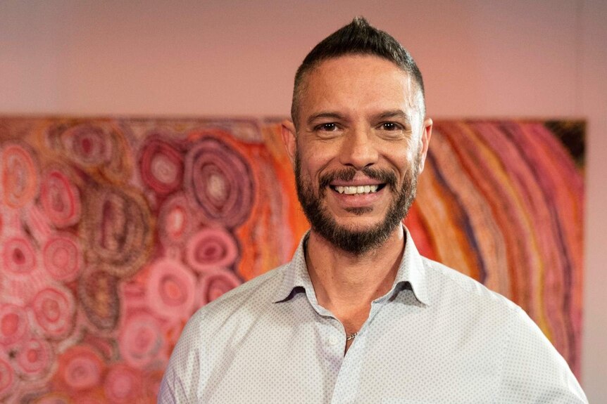 An Aboriginal man in his 40s with short dark hair and beard stands in front of an Aboriginal line and dot painting, smiling.