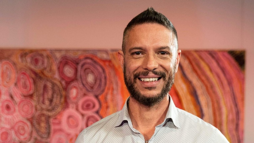 An Aboriginal man in his 40s standing in front of an Aboriginal line and dot painting and smiling.