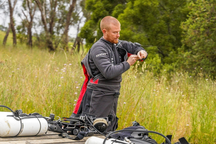 A man pulls on a wetsuit in a bushy area.