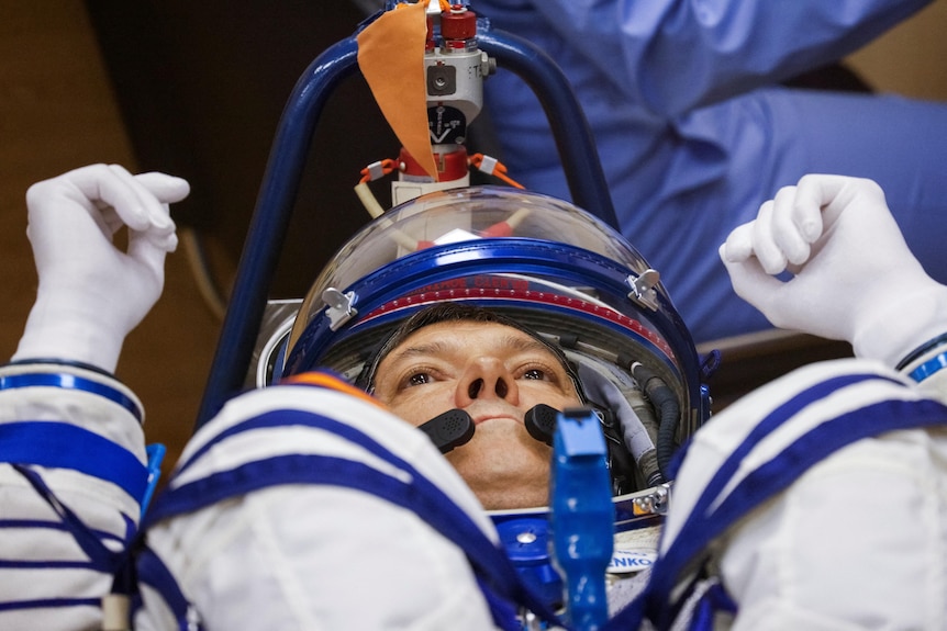 A close-up view shows a man's face in a white space suit as he lies on his back.