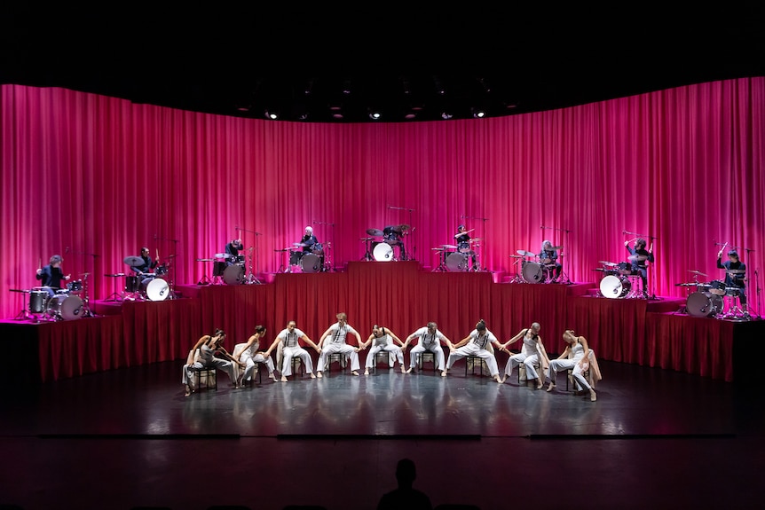 Nine drummers sit equally spaced behind kits on raised platforms. In front of them are nine dancers on chairs. 