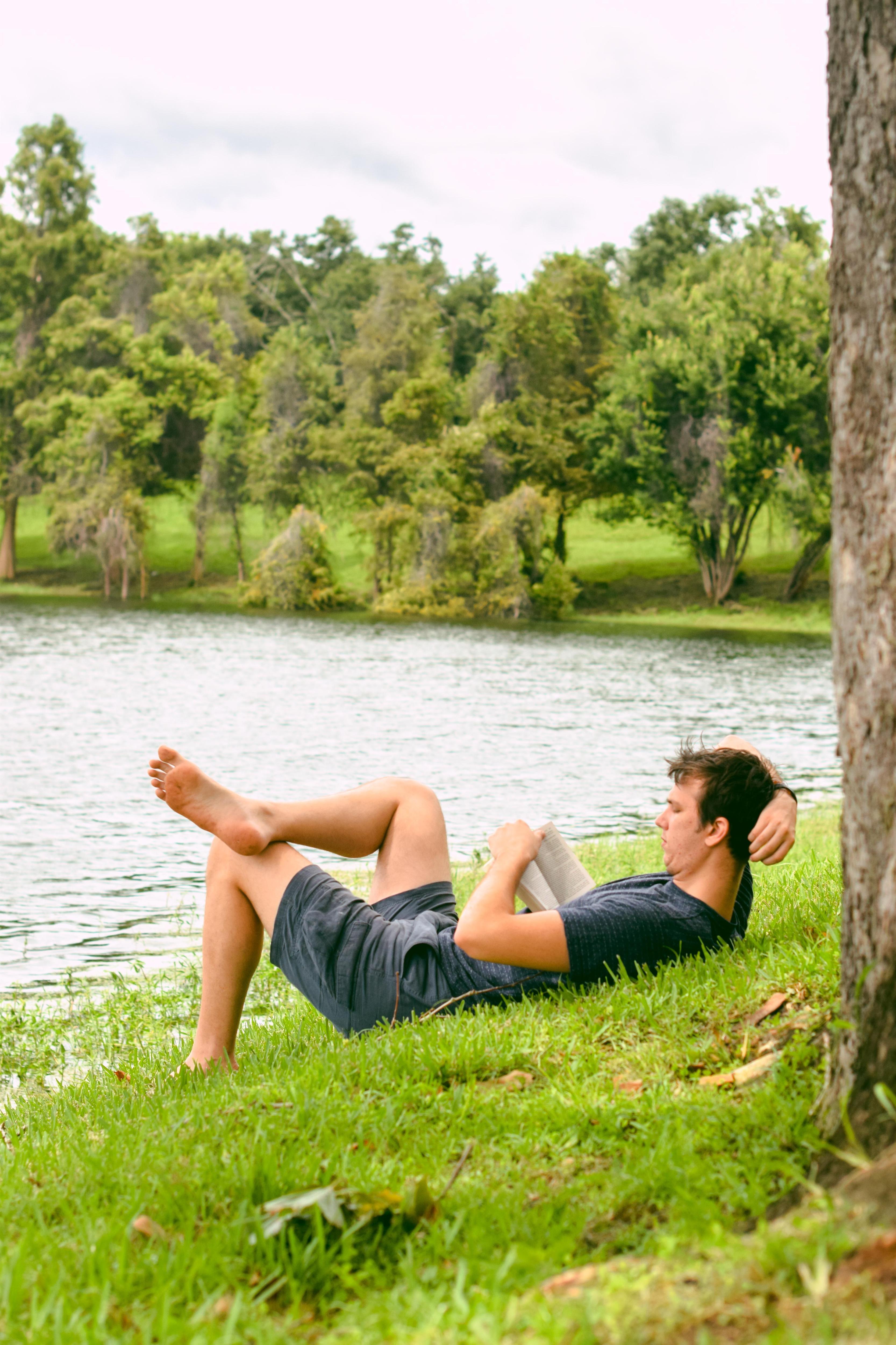 Man in shorts and t-shirt laying on grassy bank near lake reading a book.