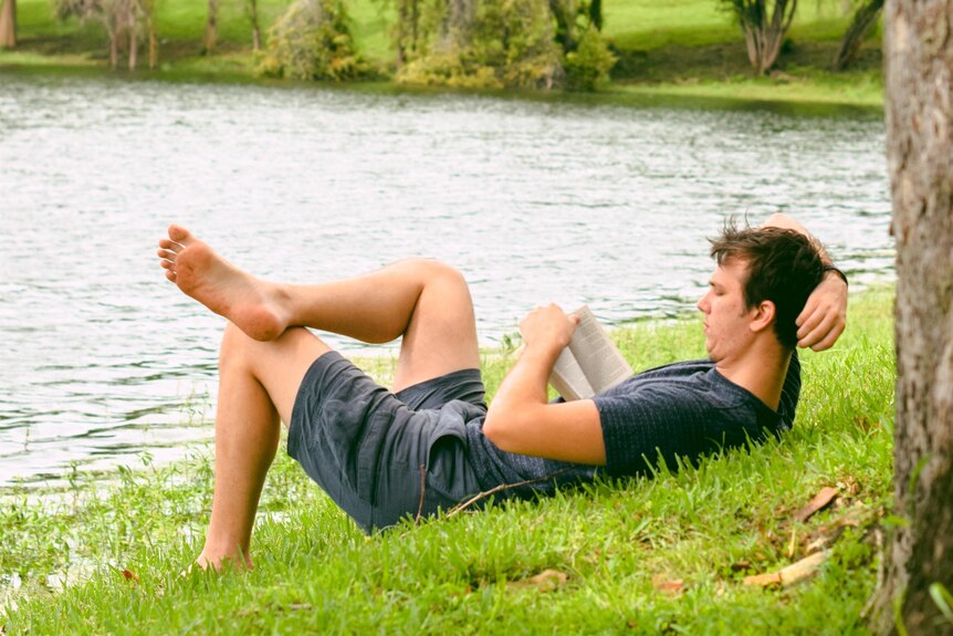 Man in shorts and t-shirt laying on grassy bank near lake reading a book.