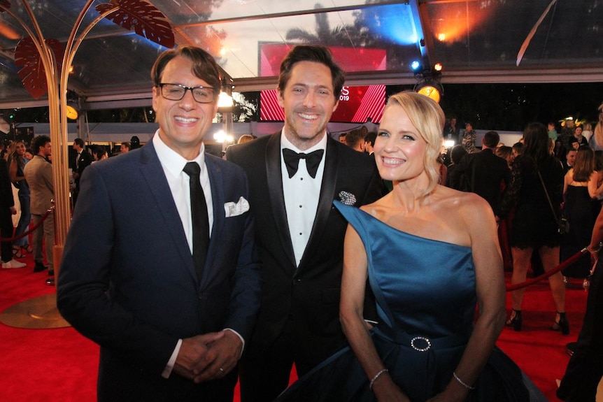 Two men in suits and a woman in a blue gown smile for the camera on a red carpet.
