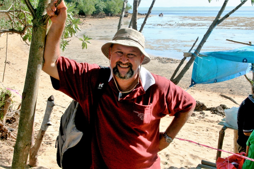 A man wearing a bucket hat and maroon t-shirt leans on a tree branch smiling with blue beach in the background
