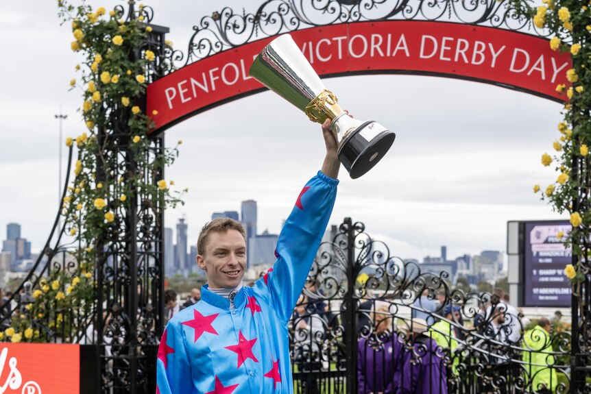 Michael Dee holds up a trophy with one hand while still wearing his pink and blue silks