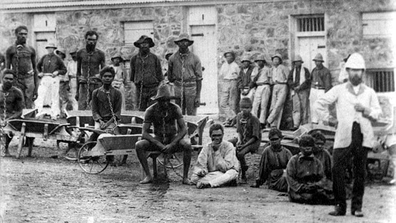 Historic black and white image of Aboriginal prisoners, many shackled, outside Fremantle prison with guards.