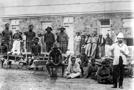 Historic black and white image of Aboriginal prisoners, many shackled, outside Fremantle prison with guards.