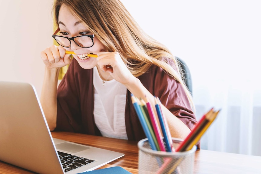 A woman bites a pencil while sitting on a chair in front of a computer during daytime.