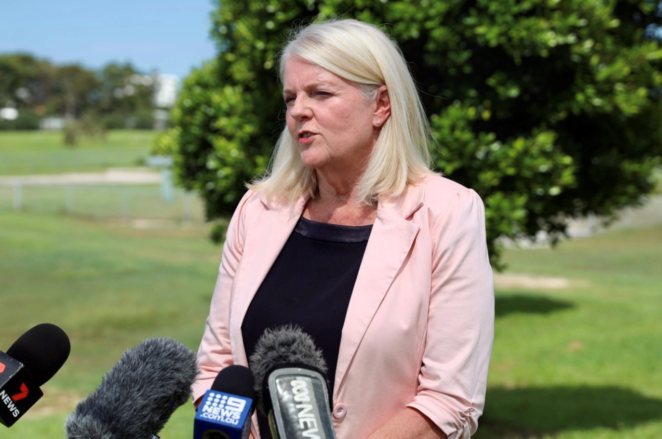 Woman with shoulder-length blinge hair, wearing oowder pink blazer and black shirt, addressing the media. 